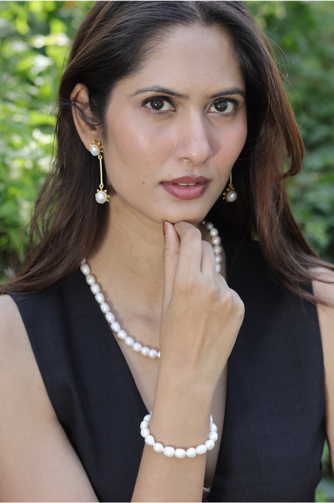 A woman with long brown hair wearing a black sleeveless top, pearl necklace, pearl bracelet, and pearl drop earrings poses outdoors, touching her chin with one hand. Green foliage is blurred in the background.