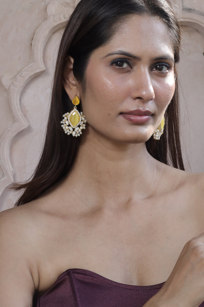 A woman with long dark hair wearing a strapless maroon top and ornate yellow and white earrings poses in front of a decorative beige wall.