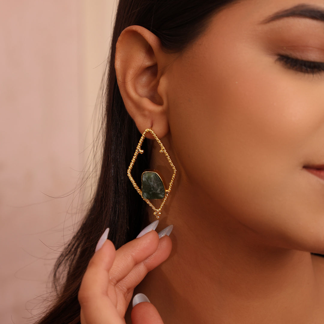 Close-up of a person wearing gold earrings with green gemstones against a neutral background