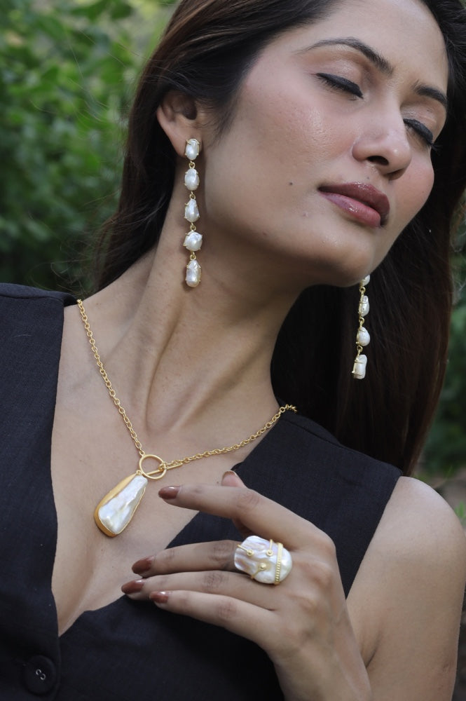 A woman with long dark hair wears pearl and gold jewelry, including dangling earrings, a pendant necklace, and a statement ring. She is dressed in a black v-neck outfit and poses outdoors with her eyes closed.